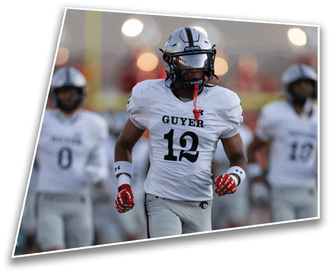 A person wearing a white football uniform with the number 12 and the word Guyer on the front is standing on a field with blurred players and lights in the background. Description generated by AI