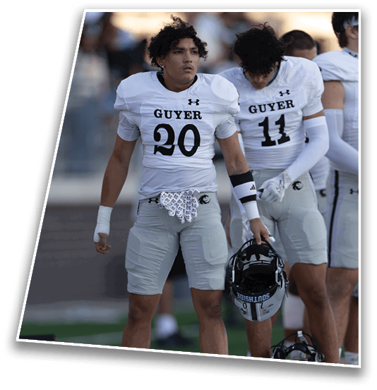 A person with curly hair wearing a white football uniform with the number 20 and the word “GUYER" stands on a field with a blurred crowd in the background. Description generated by AI