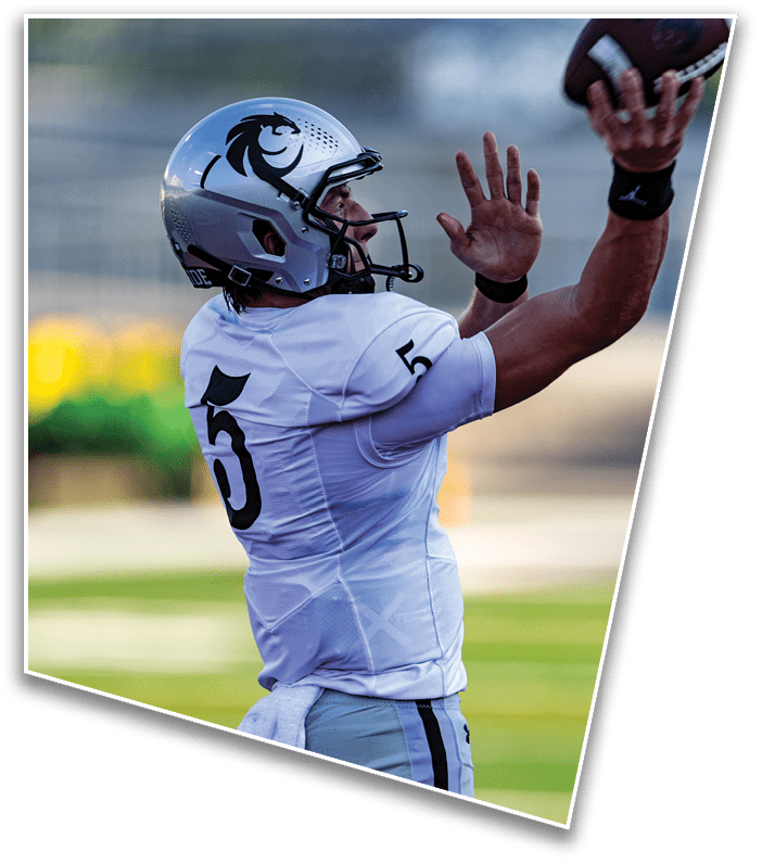 A person wearing a white football helmet with a black lion logo and a white jersey is raising one hand while looking to the side. Description generated by AI