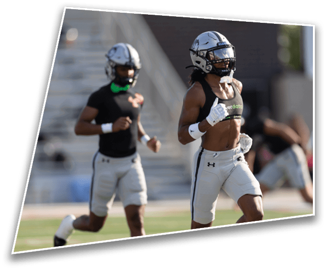 Two people wearing football helmets and dark sports uniforms are standing outdoors with a blurred background of bleachers and a building. Description generated by AI