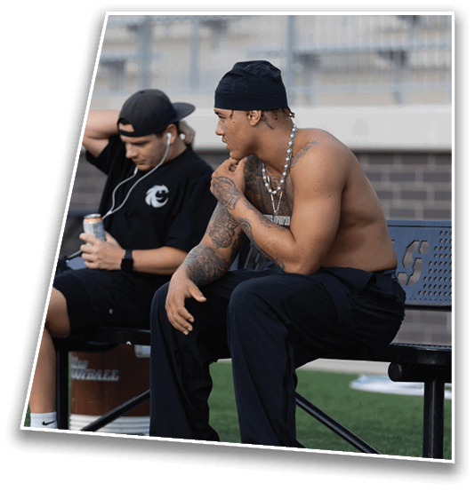 Two adults sit closely together outdoors with one wearing a black cap and earphones while the other wears a black head covering, a necklace, and has visible tattoos. Description generated by AI