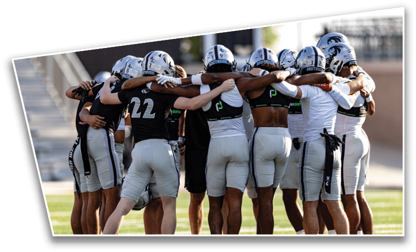 A group of football players in helmets and uniforms stand in a close huddle on a field with their arms around each other. Description generated by AI