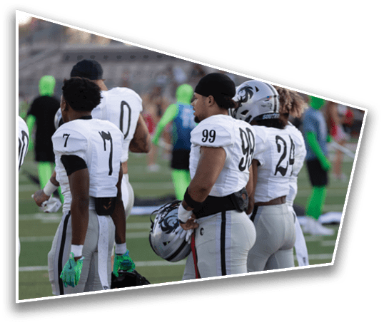 Several people wearing white football uniforms with numbers stand on a field with blurred spectators and individuals in green suits in the background. Description generated by AI