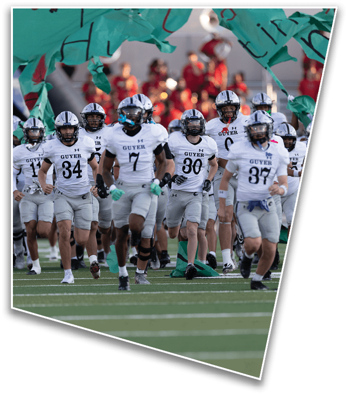 A group of football players in white and gray uniforms with helmets are running onto a field through a torn green banner. Description generated by AI