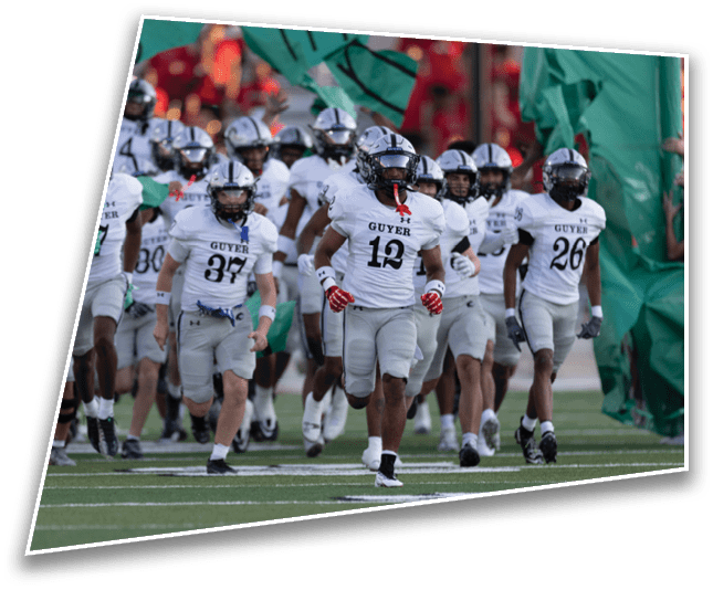 A group of football players in white uniforms with “Guyer" on their jerseys are running onto the field through a torn green banner that reads "Cardinal." Description generated by AI