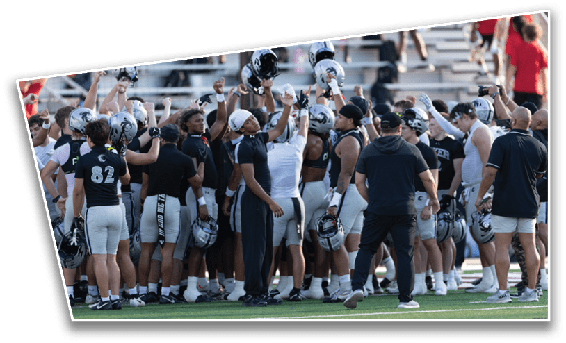 A group of football players and coaches in uniform are gathered on a field with some raising their helmets in a team huddle. Description generated by AI