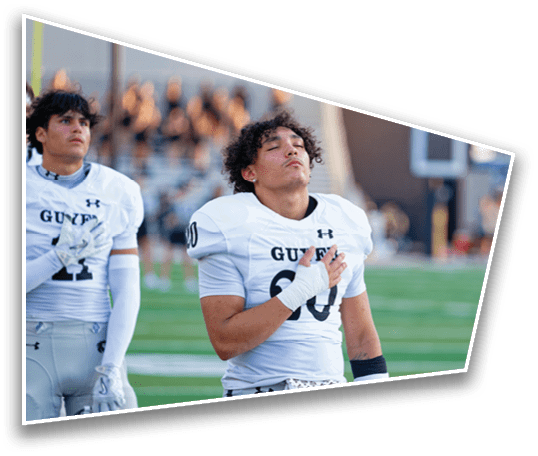 An adult wearing a white football uniform with the word “GUTIER" and the number 60 stands on a field with eyes closed and a hand placed over the chest. Description generated by AI