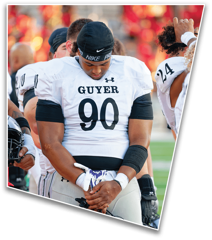 A person wearing a white football jersey with the number 90 and the word Guyer is standing with their head bowed and hands clasped, surrounded by other players on a field. Description generated by AI