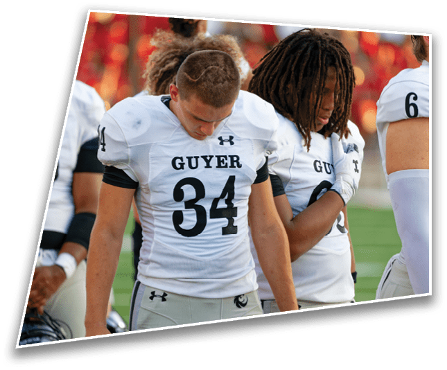 Two people wearing white football uniforms with the word “GUYER" and numbers on the front stand closely together on a field, both looking downward. Description generated by AI