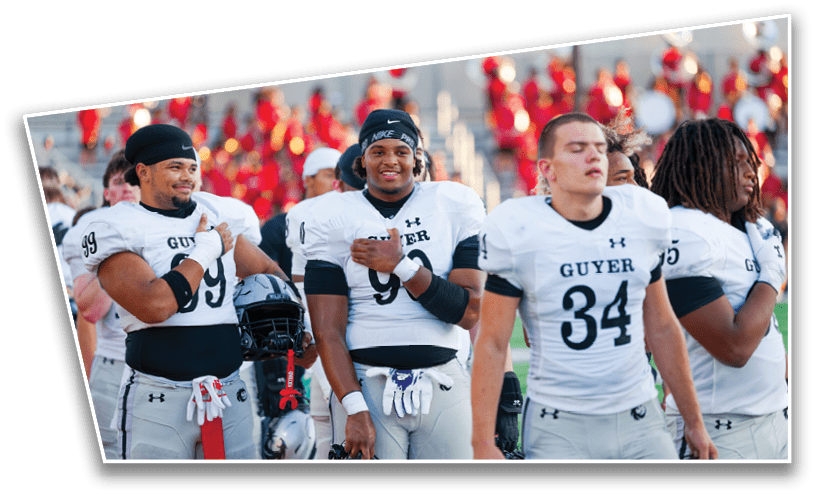 Four people wearing white football uniforms with the word “GUYER" stand side by side on a field with a blurred crowd in the background. Description generated by AI