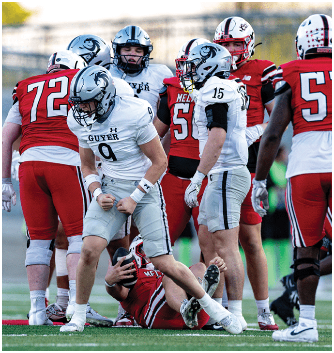 A group of people in football uniforms are on a field with one person in white celebrating while another person in red lies on the ground holding a football. Description generated by AI