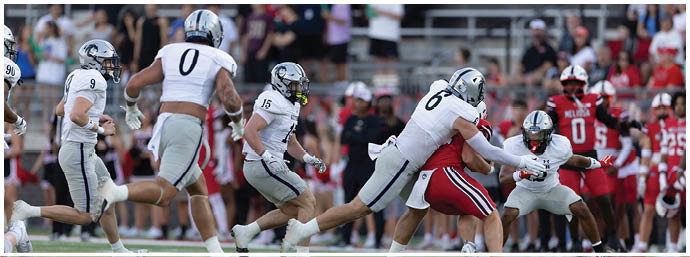 A group of people in football uniforms are actively playing on a field with one person in red being tackled by two people in white and gray uniforms. Description generated by AI