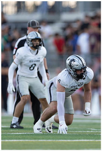 Two people in white football uniforms and one person in a red football uniform are positioned on a field during a game, with one person in white crouched and ready to move. Description generated by AI