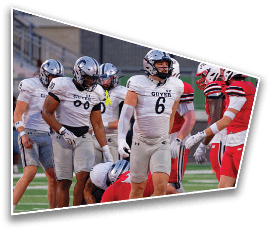 People wearing football uniforms from two different teams are gathered on a field during a game, with some standing and others crouching. Description generated by AI