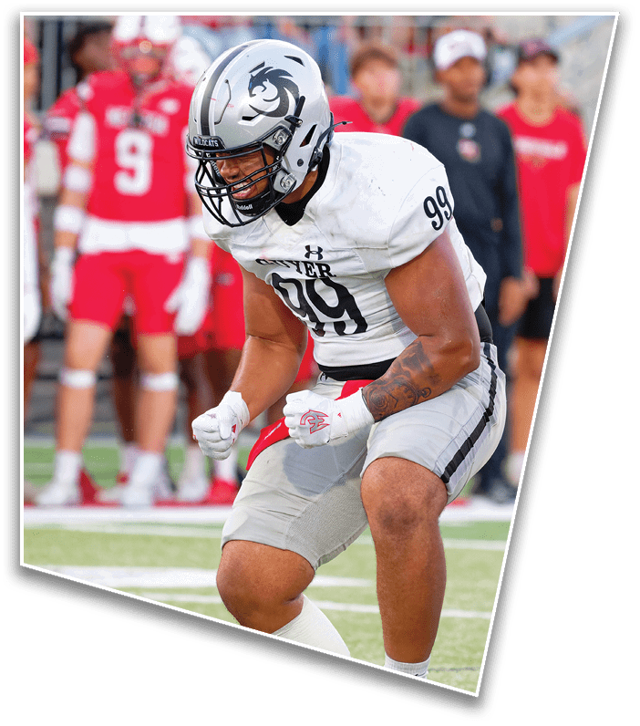 A person wearing a white football uniform and helmet with a wildcat logo is leaning forward on a field with blurred spectators in the background. Description generated by AI