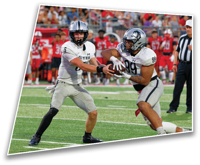 Two people in football uniforms are engaged in a handoff on a football field with a referee and a crowd in the background. Description generated by AI