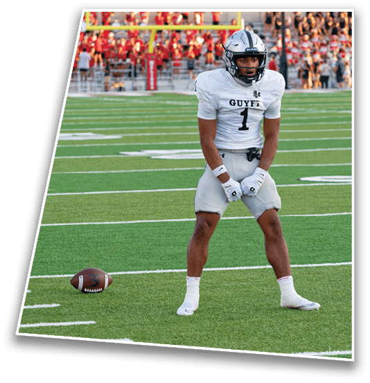 A person in a white football uniform stands on a green field near a football with a scoreboard and spectators in the background. Description generated by AI