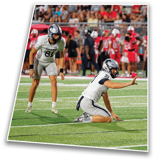 A person wearing a white and gray football uniform with a helmet is kneeling on a football field with one hand raised while other players and spectators are visible in the background. Description generated by AI