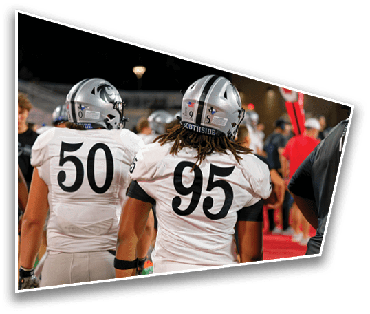 Two people wearing white football uniforms with numbers 50 and 95 stand on a field with a blurred background of other people and stadium lights. Description generated by AI