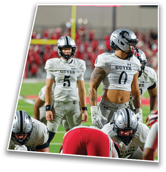 Several football players in white and gray uniforms with helmets are positioned on a field during a game, with one player standing behind others who are crouched down. Description generated by AI