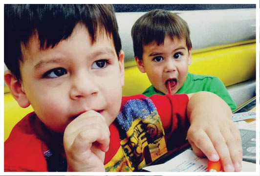 Two children are sitting at a table with one child in a red shirt looking thoughtful and the other child in a green shirt playfully sticking out their tongue. Description generated by AI