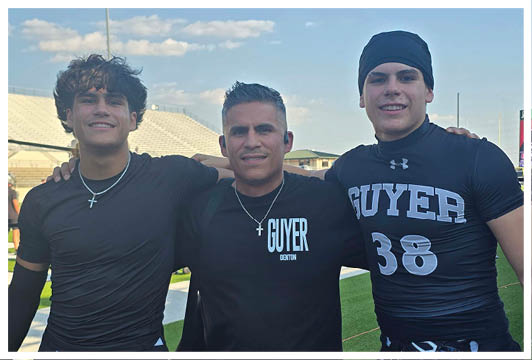 Three people wearing athletic clothing stand closely together on a sports field with one person in the center wearing a shirt that says “GUYER" and the other two wearing matching black outfits. Description generated by AI