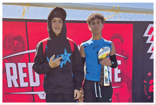 Two people stand in front of a red backdrop with text, one wearing a black hoodie and pants with blue star designs and the other wearing a blue sleeveless shirt and black pants holding a trophy. Description generated by AI
