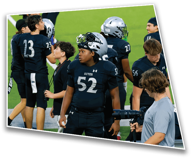 People wearing black football uniforms with helmets stand on the sidelines of a green field while one person in a gray shirt holds a camera with a large lens. Description generated by AI