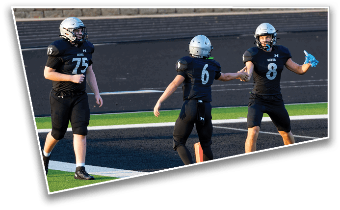 Three people in black football uniforms and helmets are on a field with one person holding a blue object and two others reaching out to each other. Description generated by AI