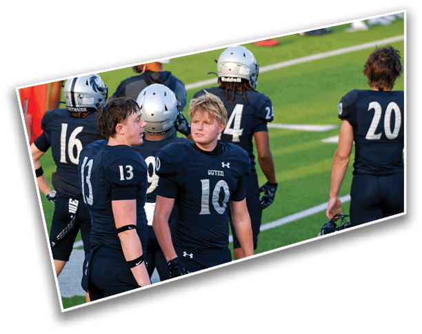 Several people wearing dark football uniforms with helmets are standing on a green field, some facing forward and others with their backs turned. Description generated by AI