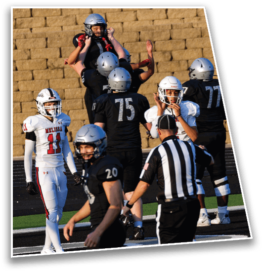 Several people wearing football helmets and uniforms are engaged in a practice or game on a field with a brick wall background. Description generated by AI