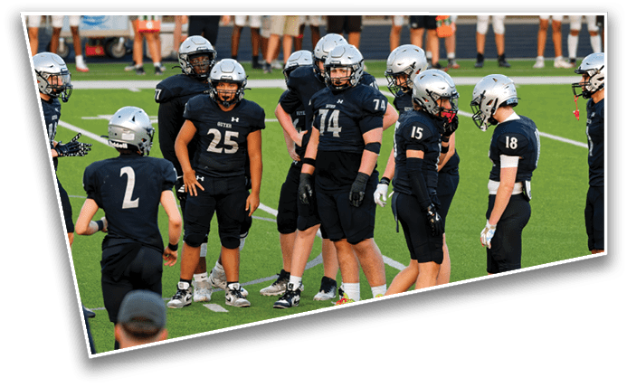 A group of people wearing black sports uniforms and silver helmets are gathered on a green field, appearing to prepare for or discuss a football play. Description generated by AI