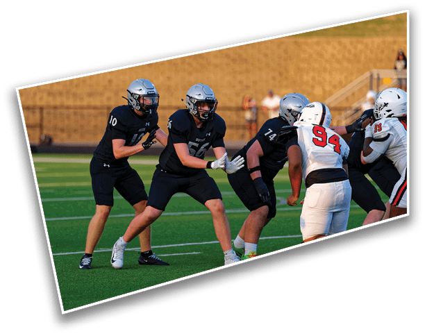 Four people wearing helmets and football uniforms are positioned on a football field, with one person in a white uniform facing three others in black uniforms. Description generated by AI