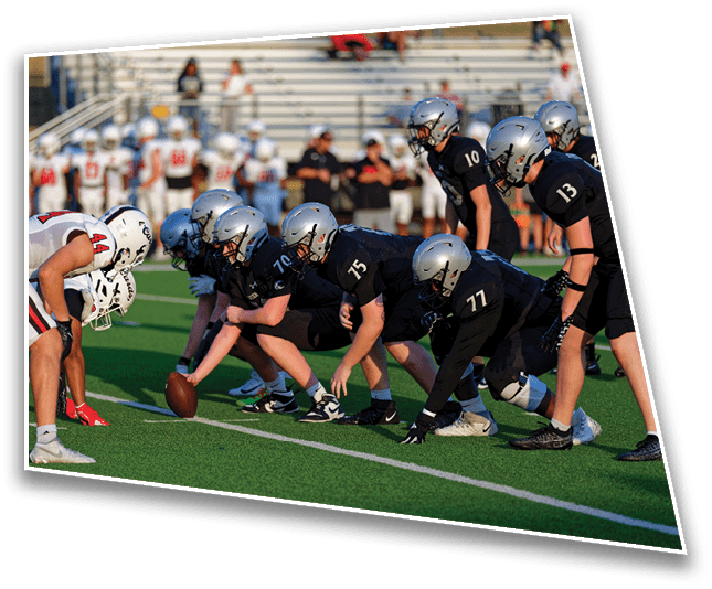 Two opposing American football teams are lined up at the line of scrimmage on a green field with empty bleachers in the background. Description generated by AI
