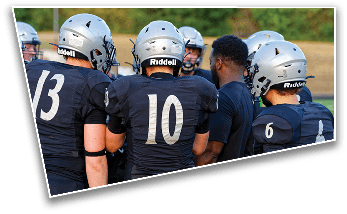 A group of people wearing football helmets and dark jerseys with numbers are gathered closely together outdoors, seemingly in discussion or strategizing. Description generated by AI