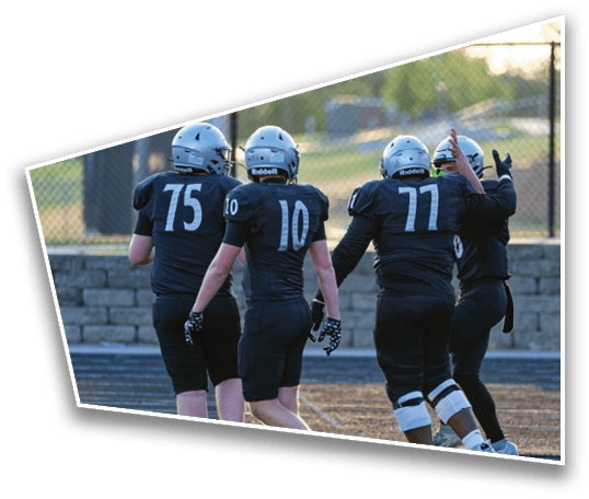 Four people in football uniforms and helmets walk together on a field near a fence, with one person raising their hands in celebration. Description generated by AI