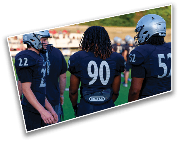 Three people wearing dark football uniforms with visible numbers stand on a grassy field, with one person wearing a helmet. Description generated by AI