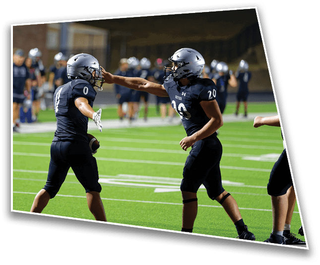 Two people in black football uniforms and helmets are on a field with one extending an arm to touch the other's helmet while holding a football. Description generated by AI