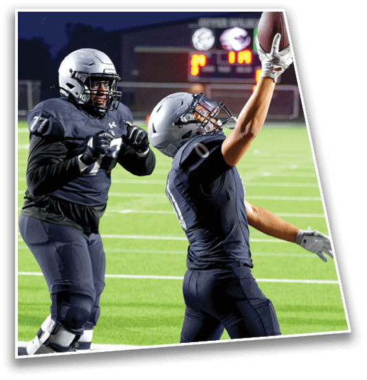 Two people in football uniforms and helmets are on a field, with one reaching up as if to catch or celebrate while the other stands nearby. Description generated by AI