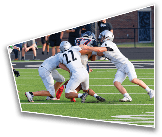 Several people in football uniforms are actively engaged in a play on a green field with a brick wall and spectators in the background. Description generated by AI