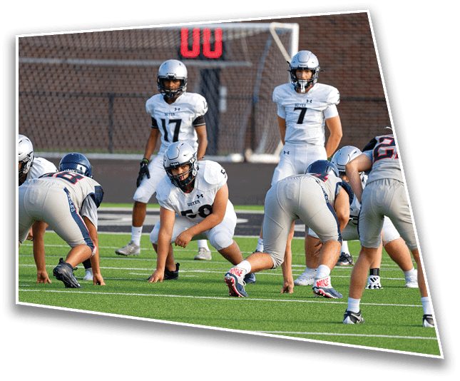 People in football uniforms are positioned on a field preparing for a play during a game. Description generated by AI