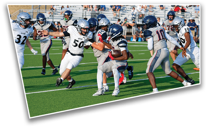 People wearing football uniforms are actively playing on a green field with one person holding the ball while others are running and attempting to tackle. Description generated by AI