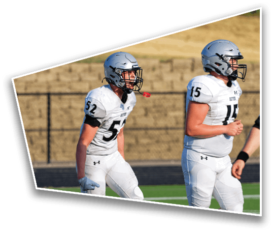 Two people wearing silver football helmets and white jerseys are on a field with a blurred natural background. Description generated by AI