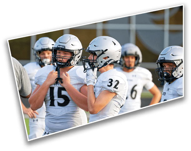 People wearing white football uniforms and helmets are gathered on a field, with two of them adjusting their helmets while others stand nearby. Description generated by AI