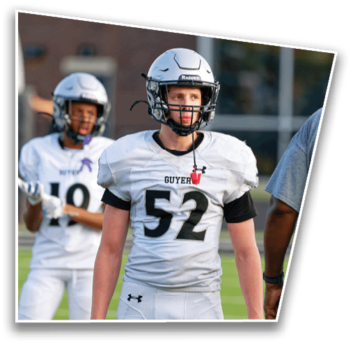 Two people wearing white football uniforms and helmets stand outdoors with a blurred building in the background. Description generated by AI