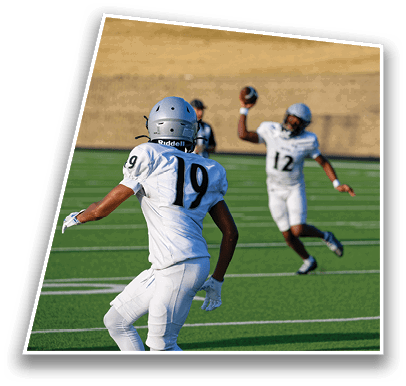 Two people in white football uniforms and helmets are on a green field with one person preparing to throw a football while the other looks on. Description generated by AI