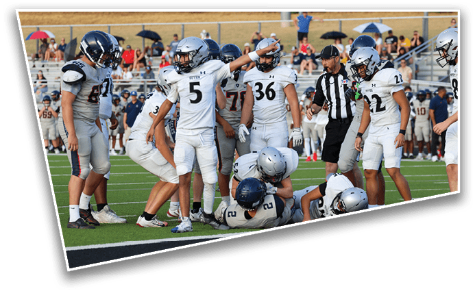 A group of football players in white and gray uniforms are gathered on a field with some players standing and others on the ground while a referee observes nearby. Description generated by AI