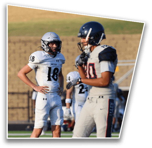 Two people in football uniforms and helmets stand on a field with one person in white and the other in gray, with a blurred background of a brick wall and grass. Description generated by AI