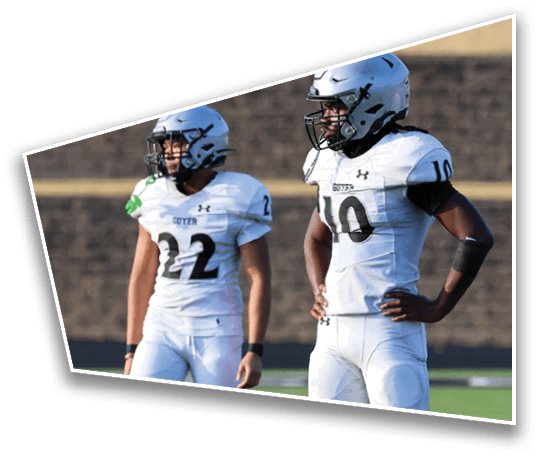 Two people in white football uniforms and helmets stand on a green field with a brick wall in the background. Description generated by AI