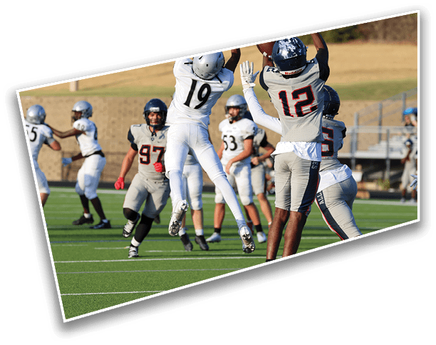 People in football uniforms are actively playing on a green field with one person jumping to catch or block a ball while others are positioned around them. Description generated by AI